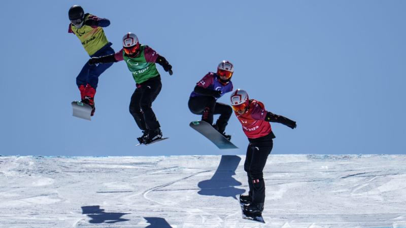 Four male snowboarders, all wearing vests that say Beijing 2022, helmets and goggles, compete at the Beijing 2022 Paralympic Games
