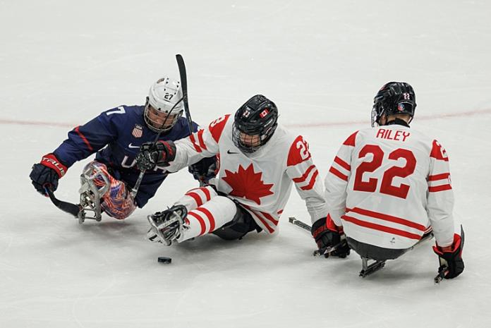 Three Para ice hockey players are trying to reach out to a hockey puck