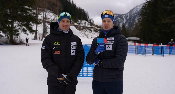 Two male athletes wearing black winter jackets are speaking outdoors in the snow
