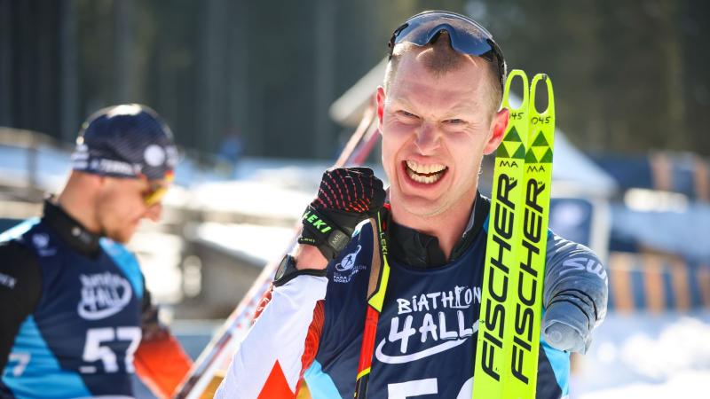 A male Para biathlon athlete smiles at the camera after competition. He is holding his skis