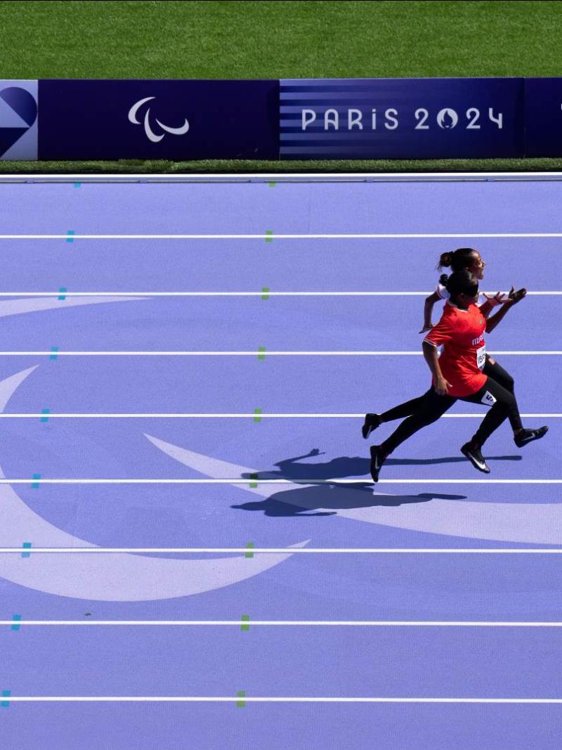 The purple track of the Paris 2024 Paralympics. There is a female athlete competing with her sighted guide