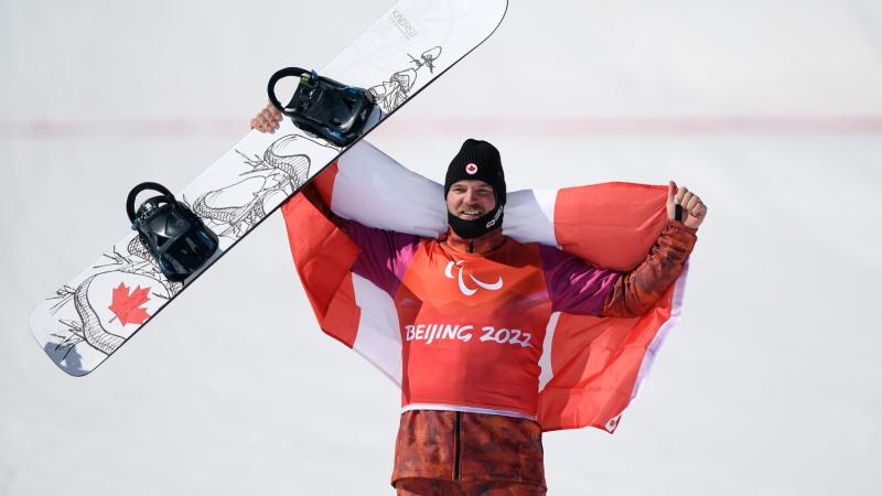 A male Para snowboarding athlete holds a snowboard and celebrates