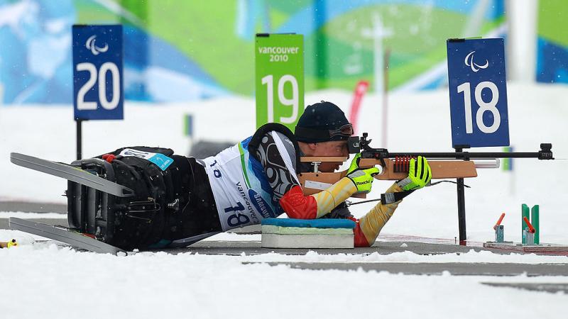 A female sit-skier is lying on the snow, aiming her rifle during a competition