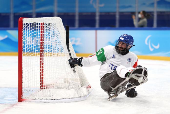 A male Para ice hockey player wearing an Italy jersey is lifting his stick on the ice in front of the net
