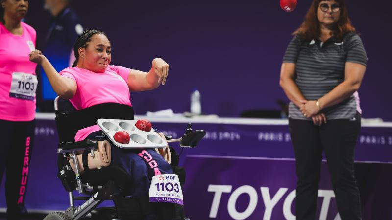 A female boccia athlete wearing a pink uniform is throwing a red boccia ball