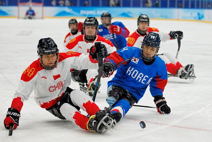 Two Para ice hockey athletes are trying to control the puck, while their teammates watch them in the background