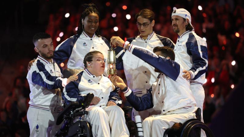 Aurelie Aubert blows out the Paralympic flame. Five athletes surround her, one of them holding a small lantern in front of her face