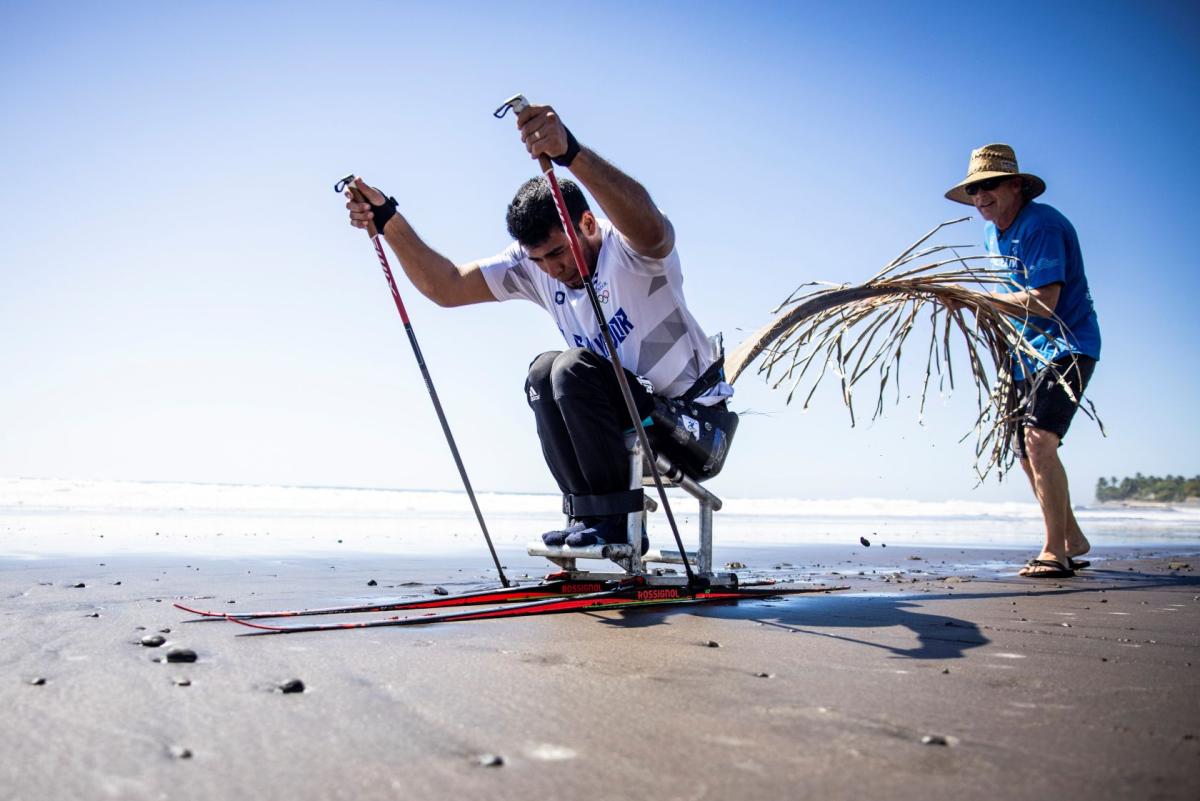 In preparation for the Milano Cortina 2026 Paralympic Winter Games, a male Para athletes trains in a sit-ski on the beach, propelling forward using two poles.