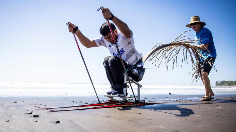 In preparation for the Milano Cortina 2026 Paralympic Winter Games, a male Para athletes trains in a sit-ski on the beach, propelling forward using two poles.