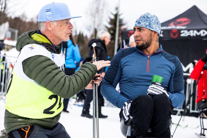 A male Para cross-country skier is talking with his coach, a male wearing a blue cap and a yellow bib