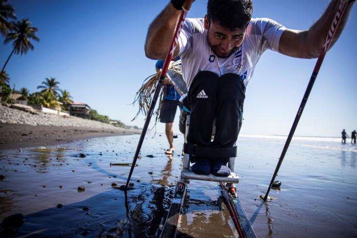 In preparation for the Milano Cortina 2026 Paralympic Winter Games, a male Para athletes trains in a sit-ski on the beach, propelling forward using two poles.