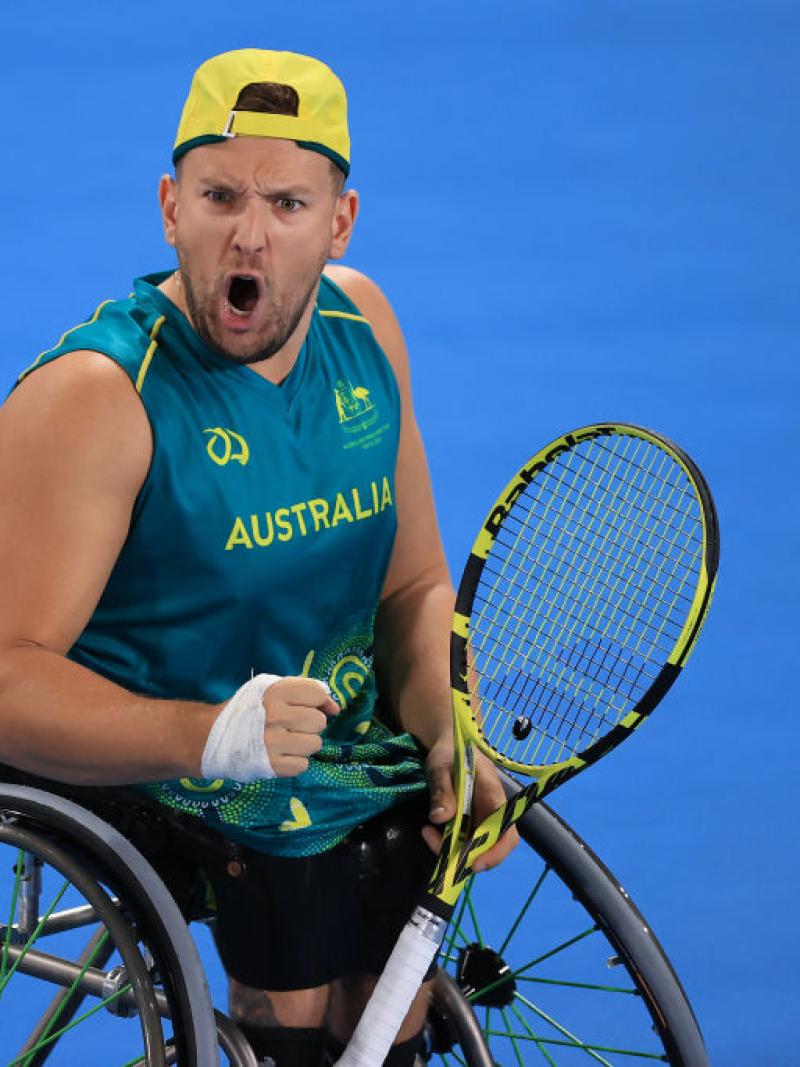 A male wheelchair tennis player yells while pumping his right fist and holding a racquet with his left hand on a blue court.