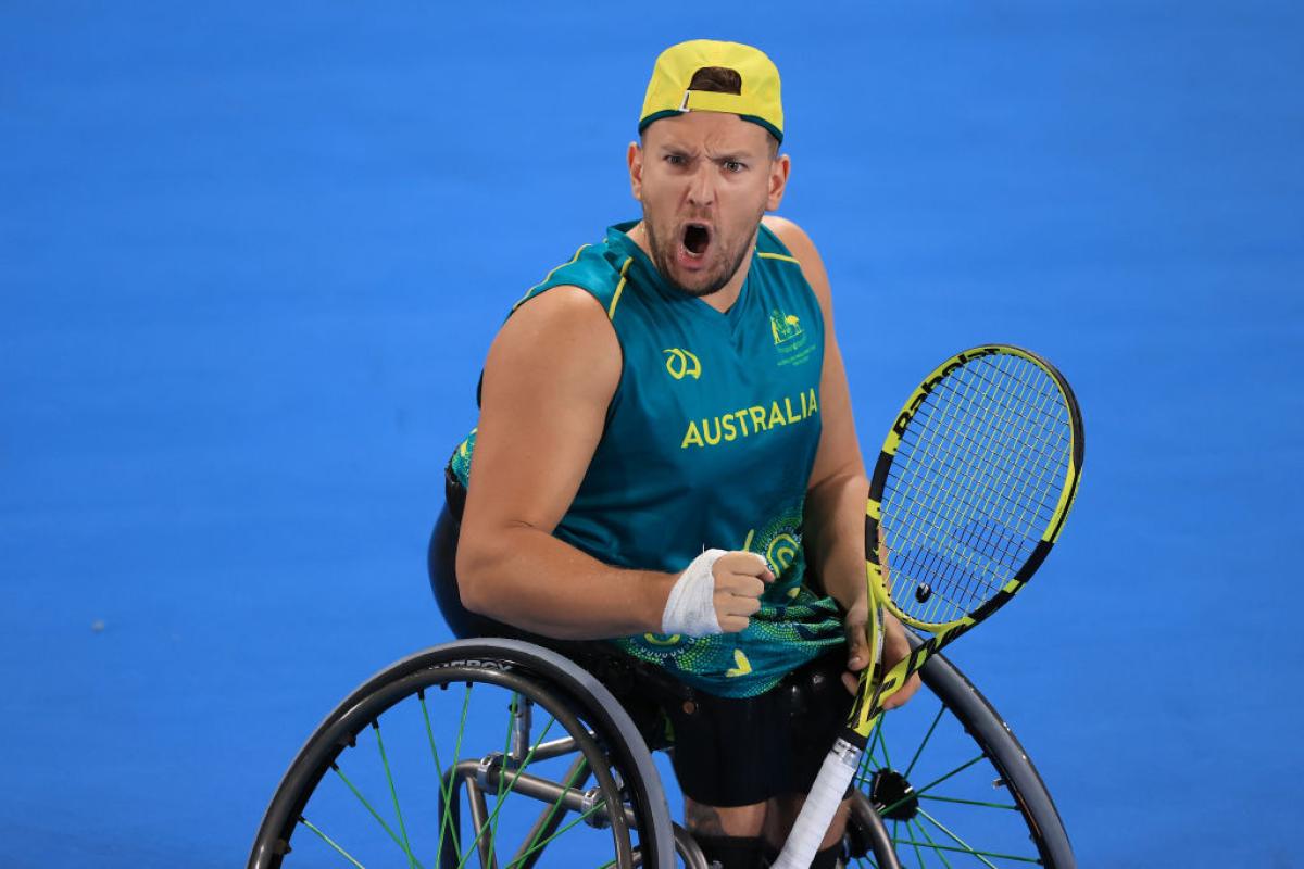 A male wheelchair tennis player yells while pumping his right fist and holding a racquet with his left hand on a blue court.