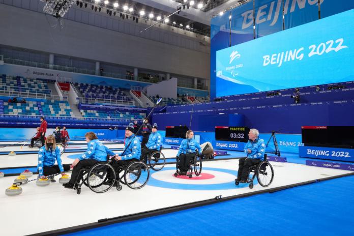Five wheelchair curling athletes and a coach dressed in blue uniform are on the ice