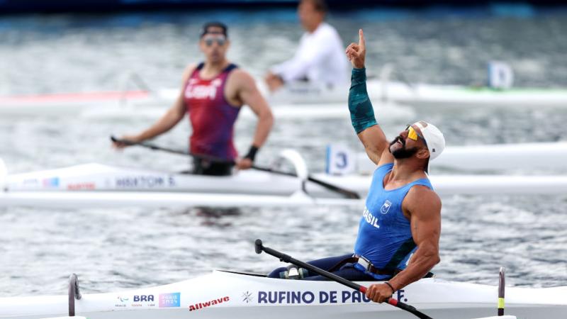 A male Para canoe athlete celebrates after winning his race - he is pointing his right index finger to the sky.