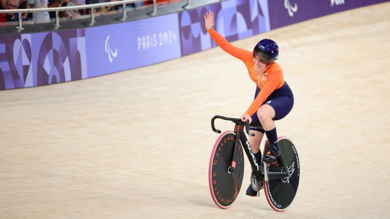 Caroline in orange with a blue helmet waves to crowd from her bike on the velodrome