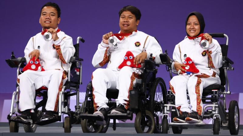 Three Indonesian boccia players in wheelchairs holding up their silver medals on the podium