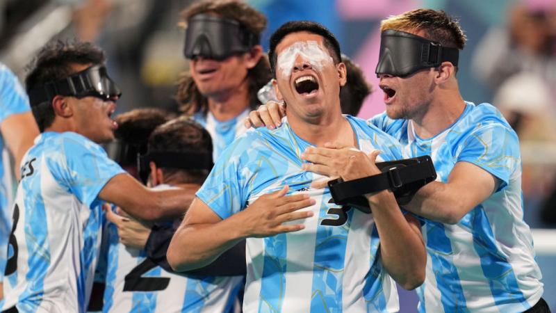 A group of men wearing Argentinian blind football uniforms and black eyeshades yell and hug each other in celebration