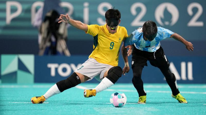 Two blind football players wearing black eyeshades, one in a yellow Brazil jersey and one in a blue and white Argentina jersey, compete for the ball
