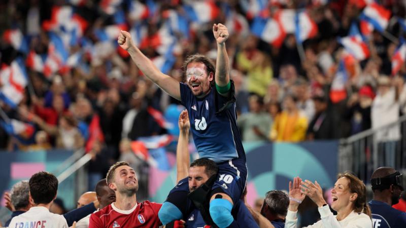 A man in a Team France football uniform with white patches over his eyes is lifted onto the shoulders of one of his teammates, as the crowd in the background waves French flags 