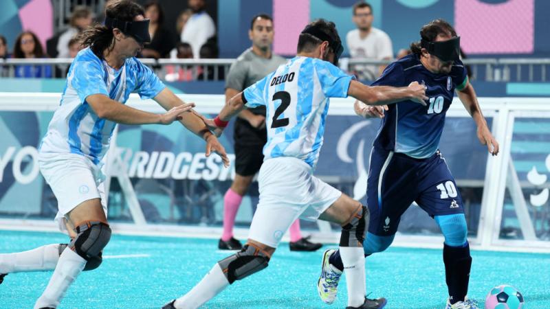 Two blind football players in Argentina uniforms and black eyeshades chase a French player, who has the ball