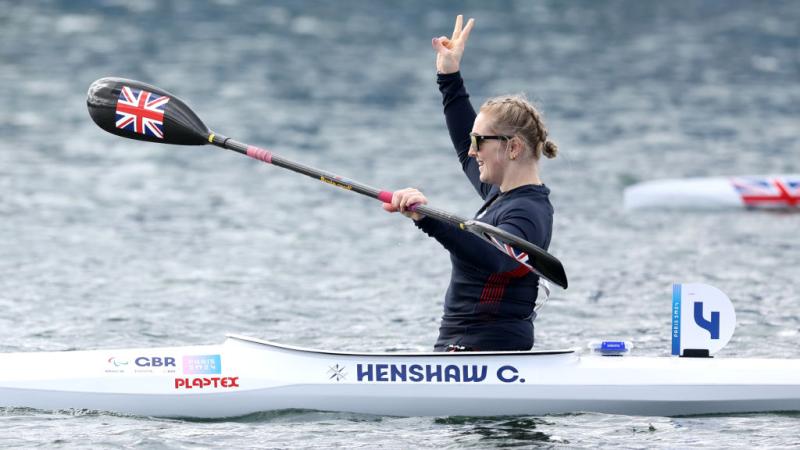 A woman in a canoe holds her oats with one hand and lifts the other hand in the air, making a peace sign