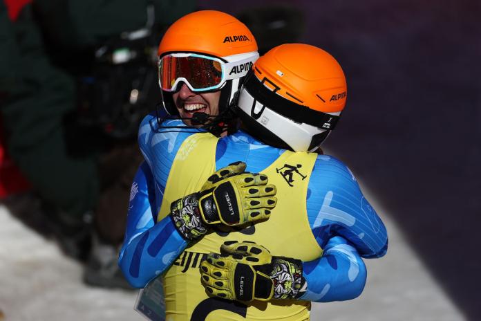 A male Paralympic alpine skiing athlete and his guide, both wearing orange headsets, are hugging