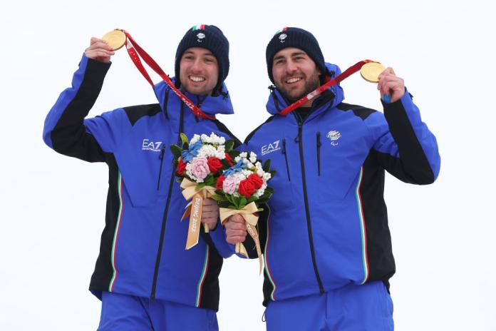 A male Paralympic alpine skiing athlete and his guide skier are celebrating on the podium at the Beijing 2022 Paralympics, posing for a photo while holding their gold medals.