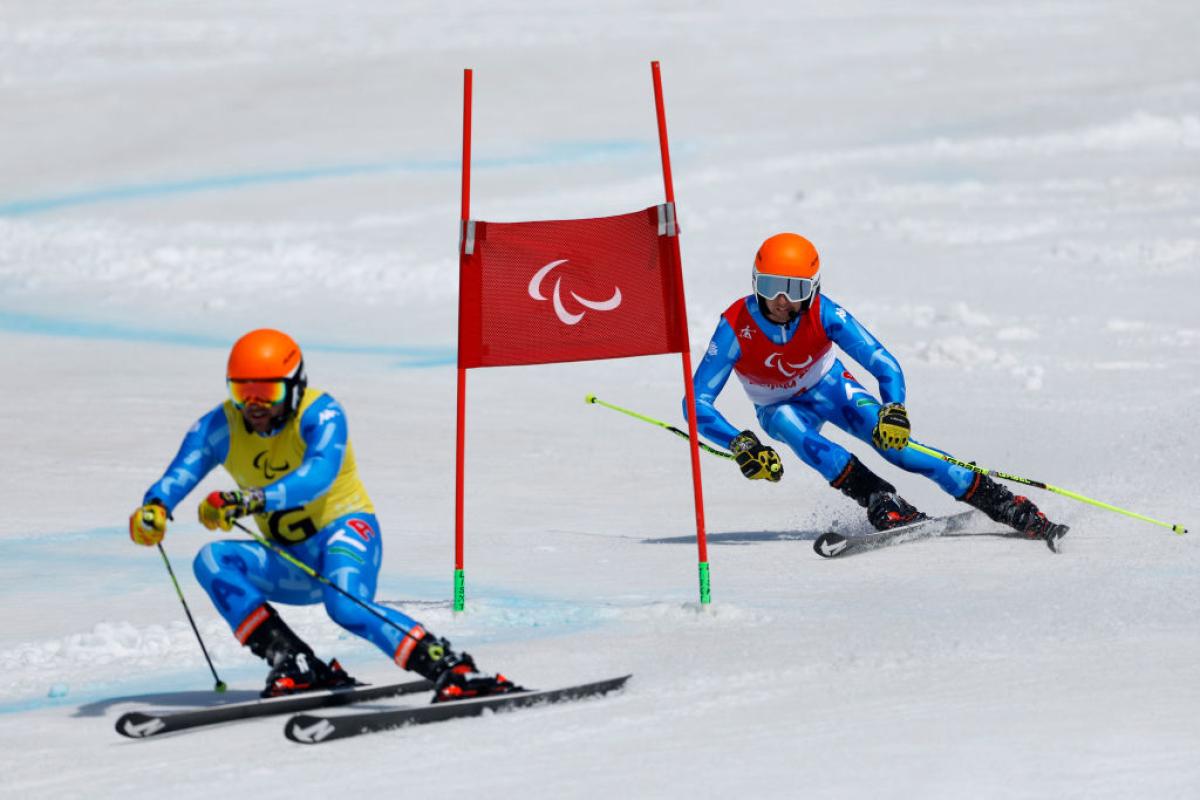 A male Paralympic alpine skiing athlete wearing a blue uniform and an orange headset is skiing behind his guide at the Beijing 2022 Paralympics.