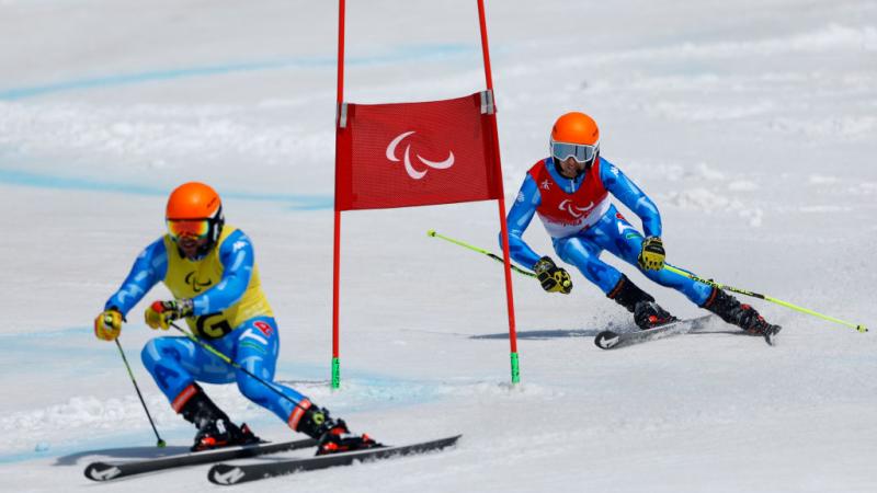 A male Paralympic alpine skiing athlete wearing a blue uniform and an orange headset is skiing behind his guide at the Beijing 2022 Paralympics.