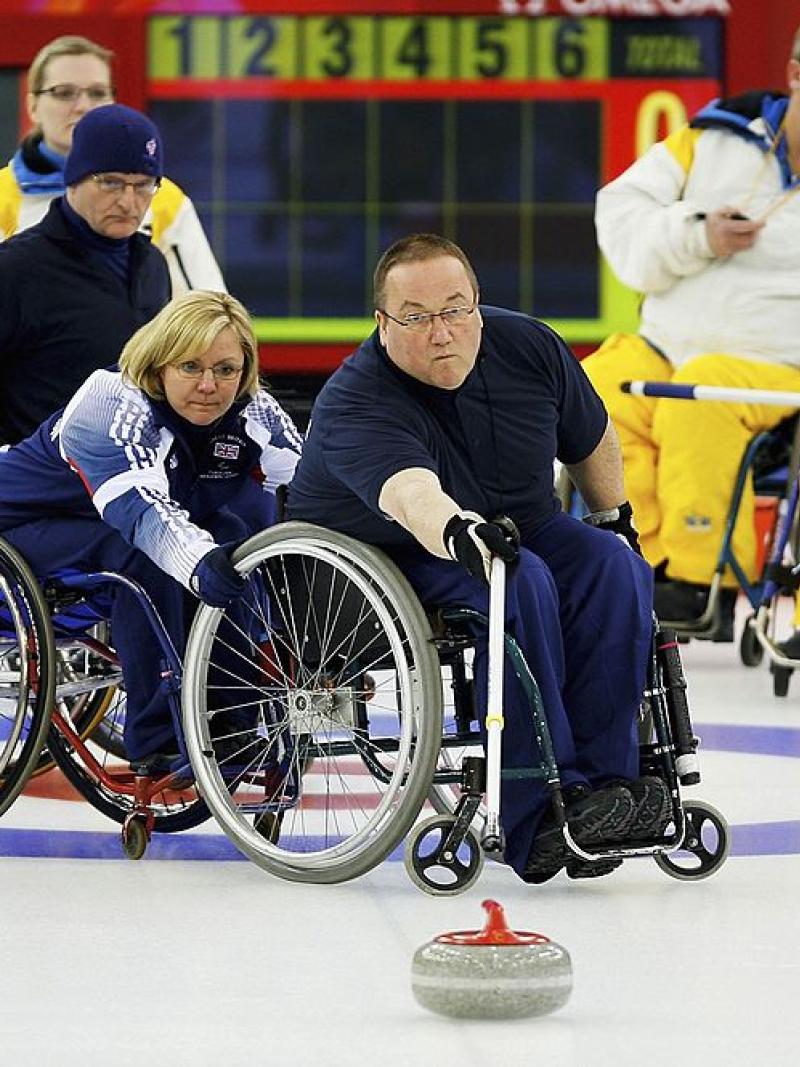 A male wheelchair curling athlete is releasing a stone with a stick. A female player is holding his wheelchair, while four athletes are watching him on the ice.