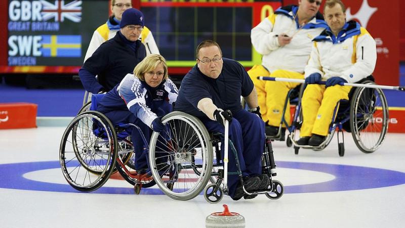 A male wheelchair curling athlete is releasing a stone with a stick. A female player is holding his wheelchair, while four athletes are watching him on the ice.