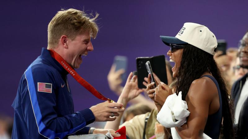 A male athlete shows his gold medal to a woman in the stands. She is holding her smartphone 