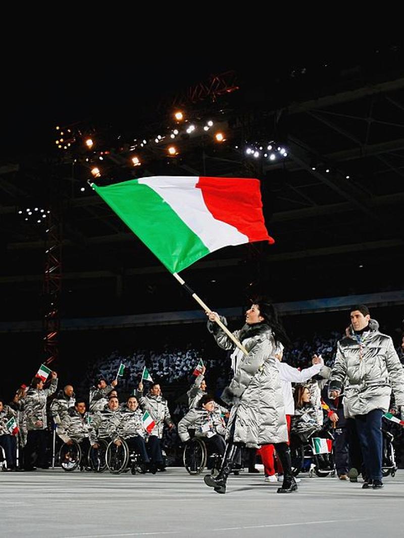 A female athlete wearing a gray winter jacket is leading the Italian delegation at the Torino 2006 Opening Ceremony by carrying the Italian flag