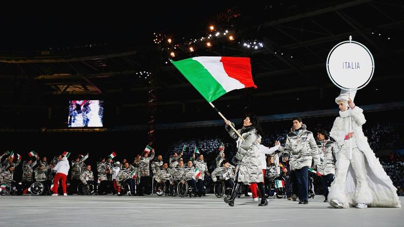 A female athlete wearing a gray winter jacket is leading the Italian delegation at the Torino 2006 Opening Ceremony by carrying the Italian flag