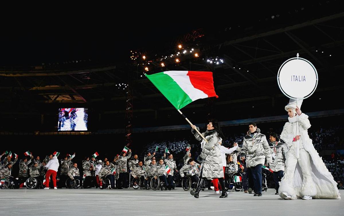 A female athlete wearing a gray winter jacket is leading the Italian delegation at the Torino 2006 Opening Ceremony by carrying the Italian flag