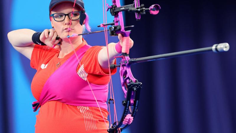 A female Para archer who is wearing a red T-shirt is aiming her bow while seven months pregnant