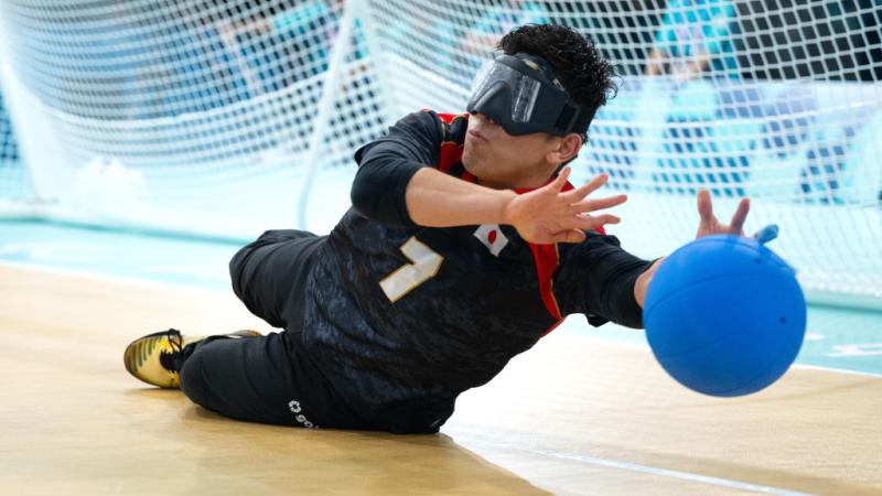 A male goalball player, wearing eye shades, is blocking a blue ball in front of the goal