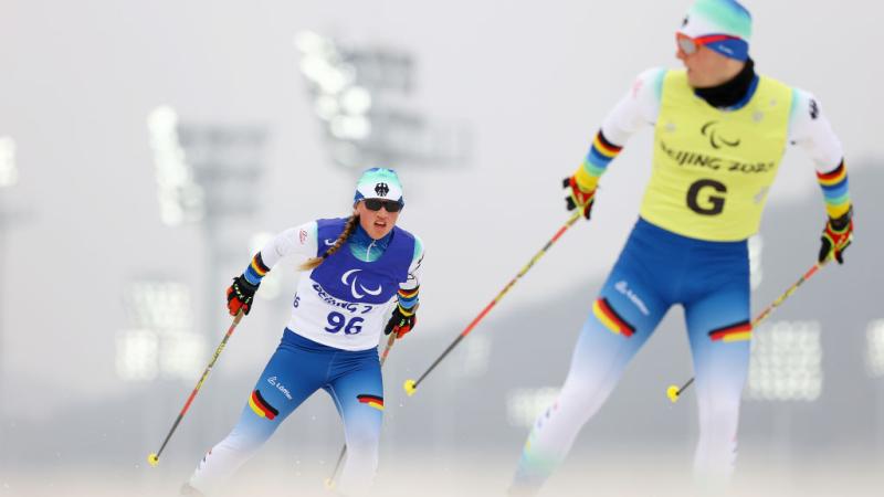 A female skier is competing behind a sighted male guide, who is looking back at her