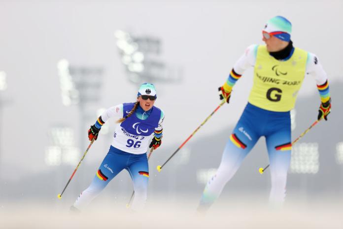 A female skier is competing behind a sighted male guide, who is looking back at her