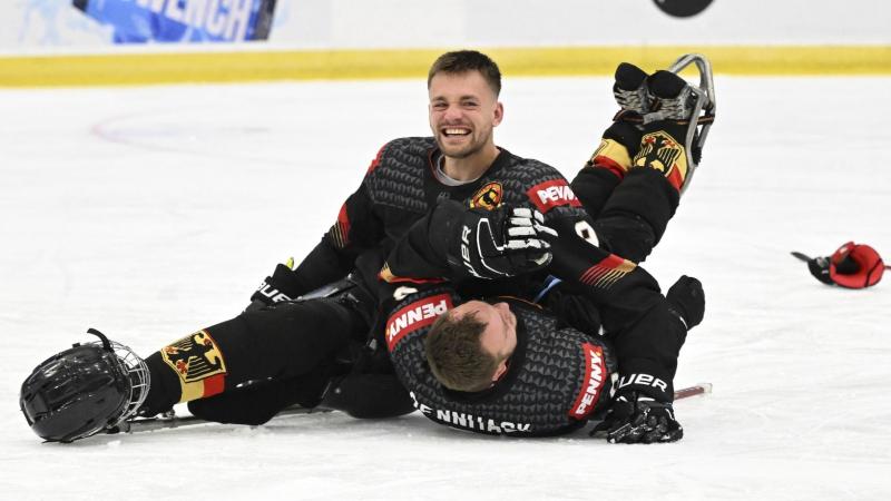 A male Para ice hockey player smiles on the rink, celebrating with his teammate who is lying on the ice