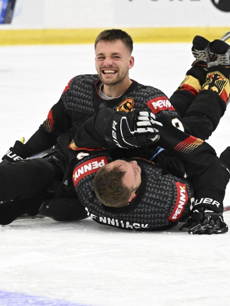 A male Para ice hockey player smiles on the rink, celebrating with his teammate who is lying on the ice