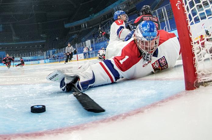 A male Para ice hockey player reaches out with a stick to block a goal in front of the net
