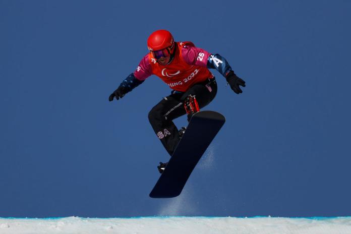 A male Para snowboarder wearing a red prosthetic leg and red helmet is making a jump during a competition at the Beijing 2022 Paralympic Winter Games.