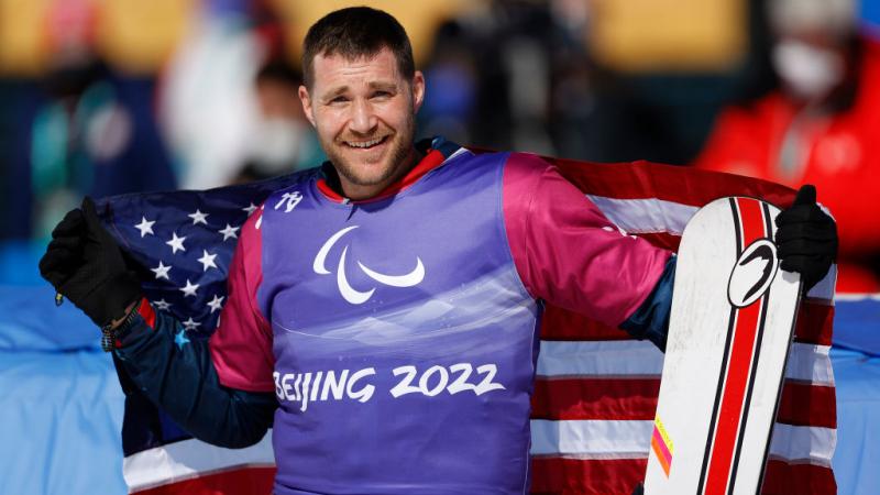 A male Para snowboarder wearing a purple bib is smiling while holding the USA flag and a snowboard