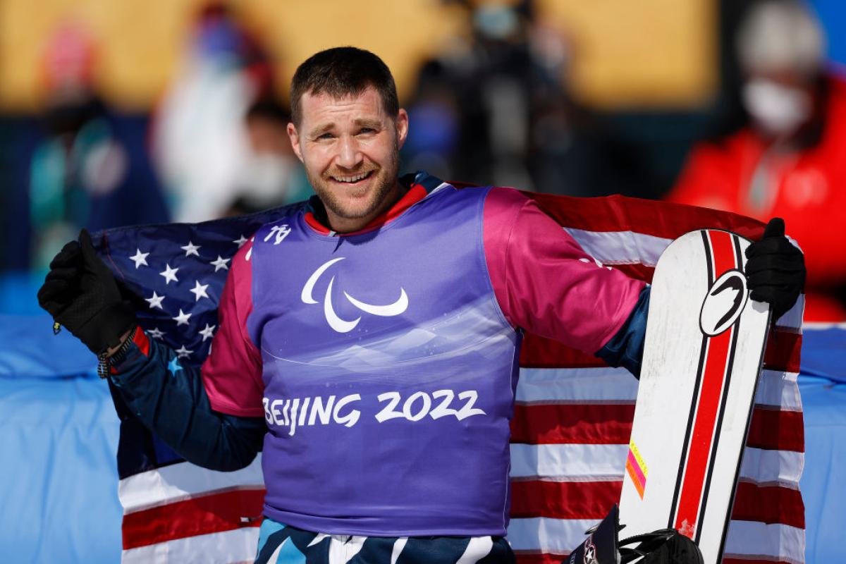 A male Para snowboarder wearing a purple bib is smiling while holding the USA flag and a snowboard