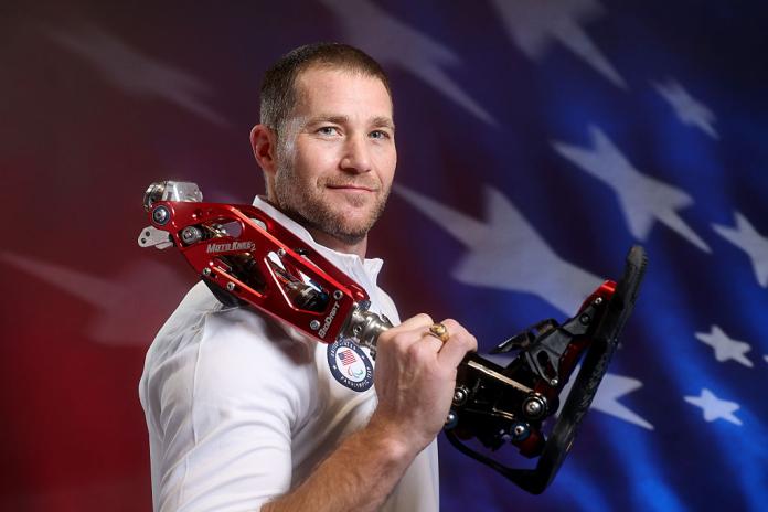 A male Para athlete is posing in front of a screen projecting the USA map while holding a red prosthetic leg 