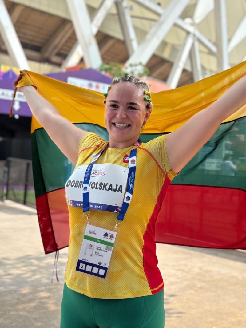 A female athlete wearing a green, yellow and red athletic gear is holding the Lithuanian flag in front of a stadium