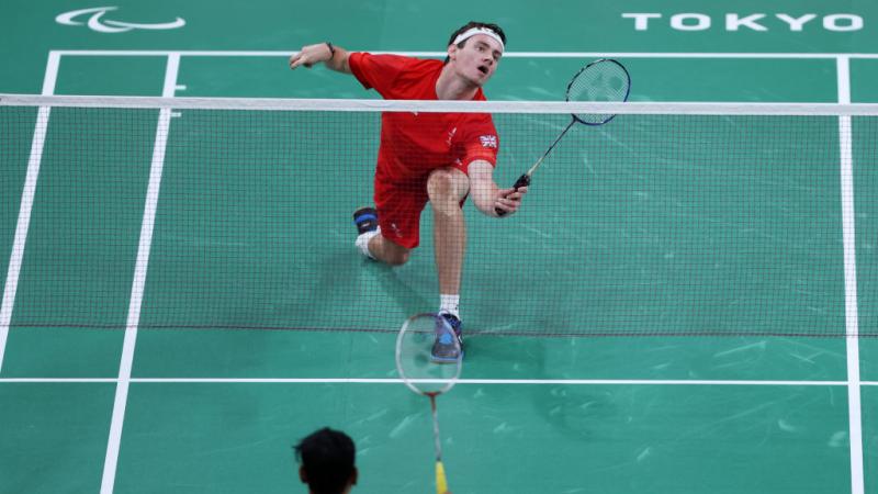 A Para badminton match between two male athletes at Tokyo 2020. A player in red uniform prepares to return the shuttle near the net.