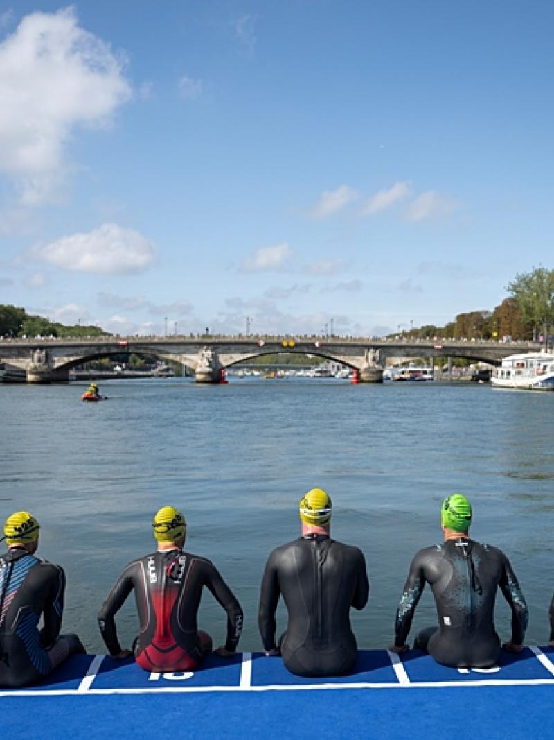 Six Para triathlon athletes are preparing for the swimming segment. The photo captures their back, sitting in front of a river